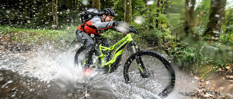 Ein Radfahrer fährt mit zugekniffenen Augen durch ein Bachbett im Wald. Das Wasser spritzt in alle Richtungen.