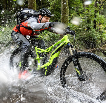 Ein Radfahrer fährt mit zugekniffenen Augen durch ein Bachbett im Wald. Das Wasser spritzt in alle Richtungen.