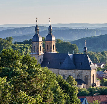 Zwischen bewaldeten Hügeln erhebt sich die imposante barocke Kirche aus Sandstein und schwarzem Schieferdach.