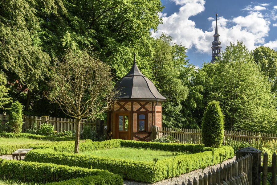 Ein Garten mit Buxbäumchen eingegrenzt und eine barocke Laube mit schwarzem Dach.