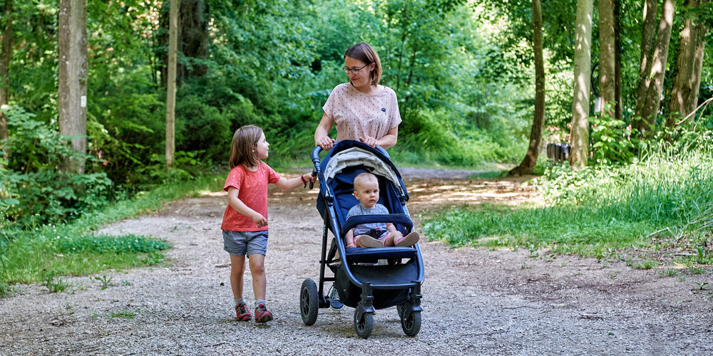 Eine Mutter mit zwei Kinder im Wald auf einem breiten Weg. Ein Kind sitzt im Kinderwagen, das andere läuft daneben und blickt zur Mutter.