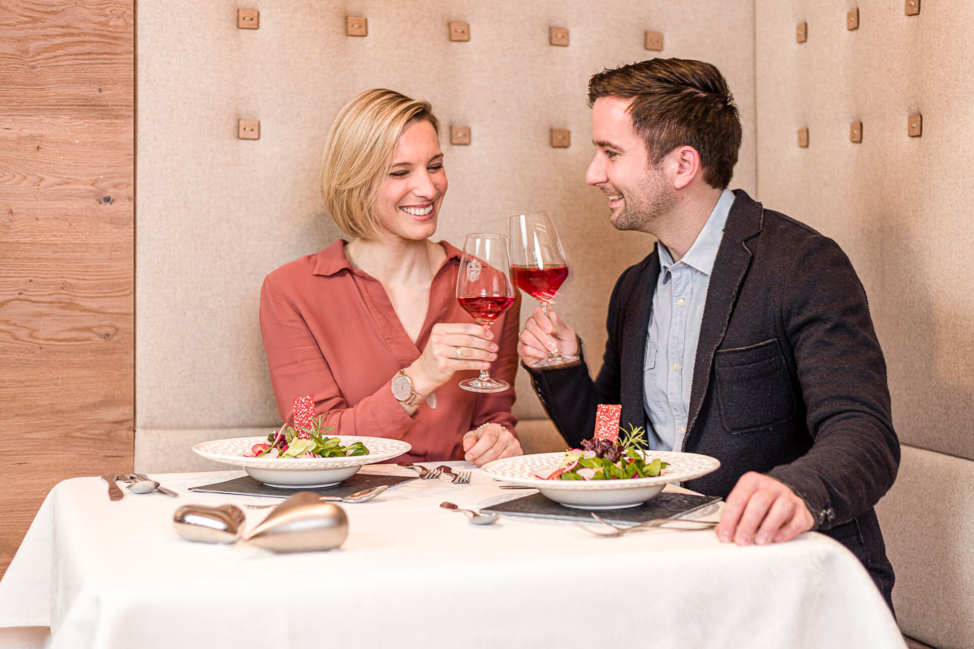 Ein Paar sitzt lächelnd an einem Tisch und prostet sich mit den Weingläsern zu. Auf dem Tisch stehen zwei Teller mit Salat.