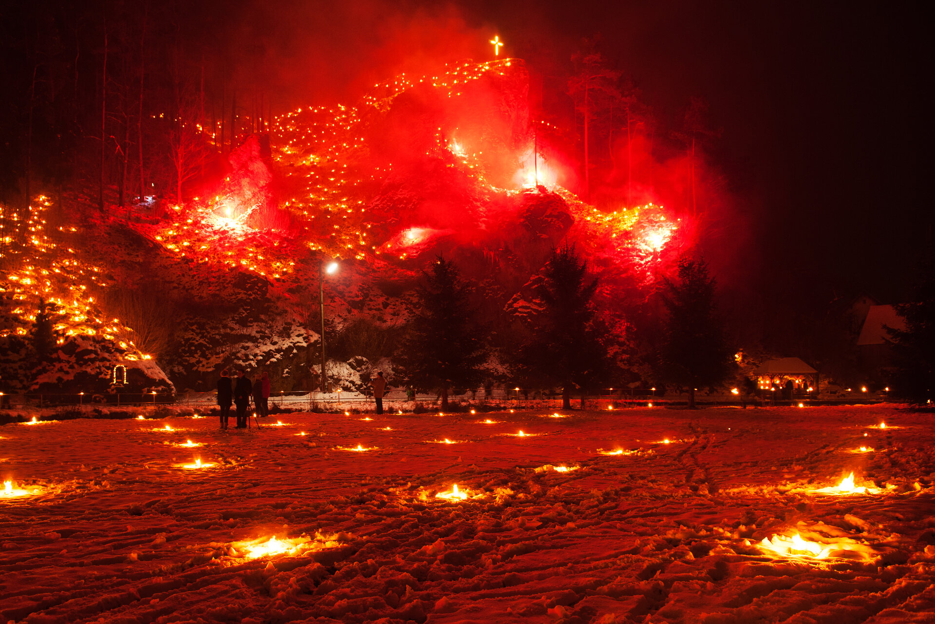 Im Vordergrund stehen Menschen auf einer schneebedeckten Wiese, in der kleine Feuer brennen. Im Hintergrund erhebt sich ein felsiger Berg, auf dem rote Feuer und eine Kreuz leuchten. 