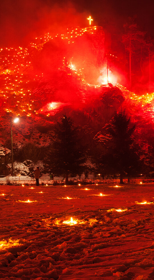 Im Vordergrund stehen Menschen auf einer schneebedeckten Wiese, in der kleine Feuer brennen. Im Hintergrund erhebt sich ein felsiger Berg, auf dem rote Feuer und eine Kreuz leuchten. 