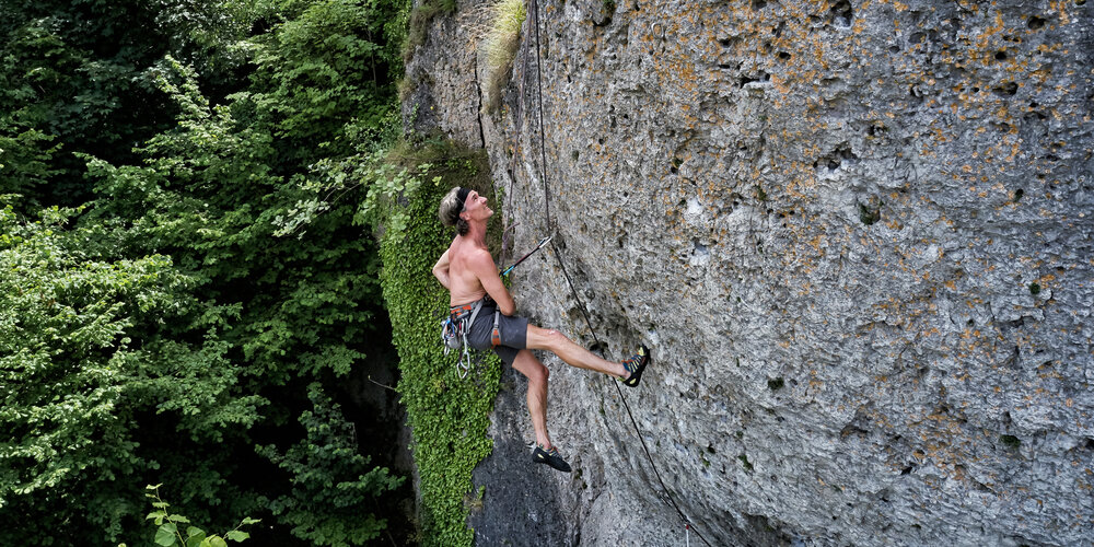 An einem großen, grauen Felsblock hängt ein Kletterer, der gerade Pause macht und am Fels nach oben blickt.