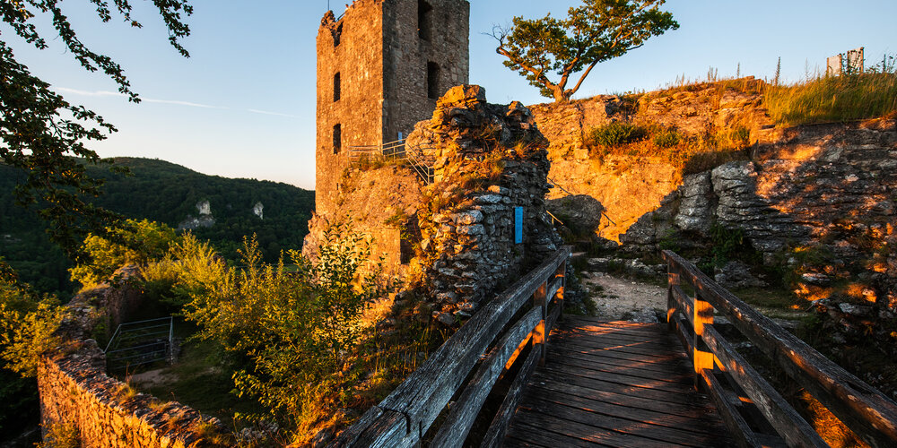 Im Vordergrund führt eine hölzerne Brücke zur dahinterliegenden Burg, die in warmen Sonnenstrahlen leuchtet.