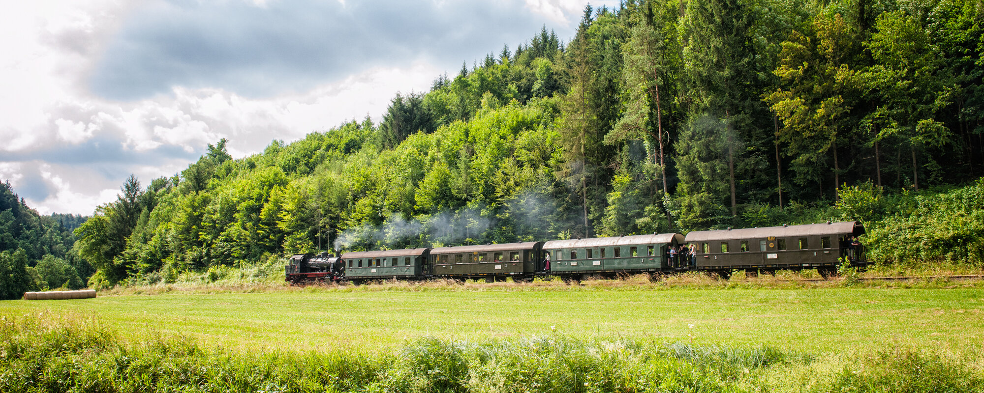 Eine alte, dampfende Eisenbahn mit vier Waggons fährt vor einem Wald. Im Vordergrund ist ein Fluss zu sehen.
