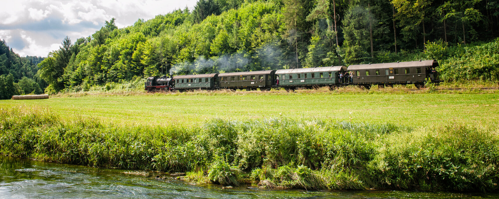 Eine alte, dampfende Eisenbahn mit vier Waggons fährt vor einem Wald. Im Vordergrund ist ein Fluss zu sehen.