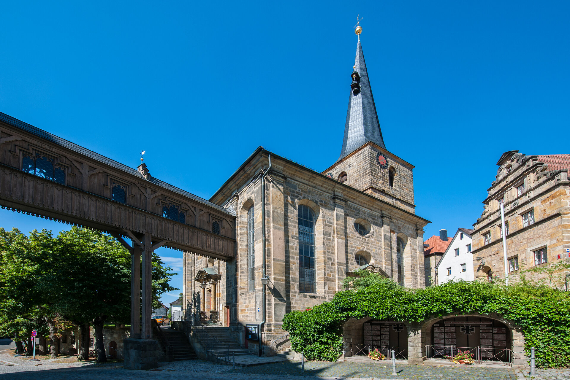 Eine Gebäudebrücke aus Holz führt von links kommend zu einer Kirche mit einem spitzen, schiefergedeckten Turm. 