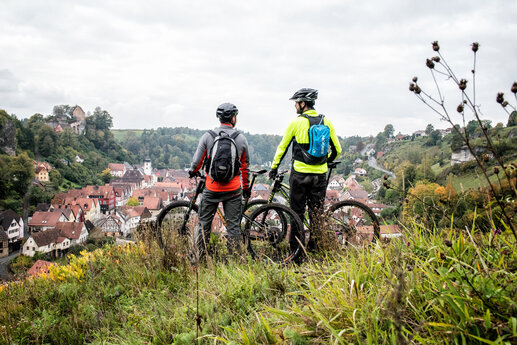 Zwei Männer von hinten stehen mit ihren Mountainbikes über dem Ort und blicken hinab.