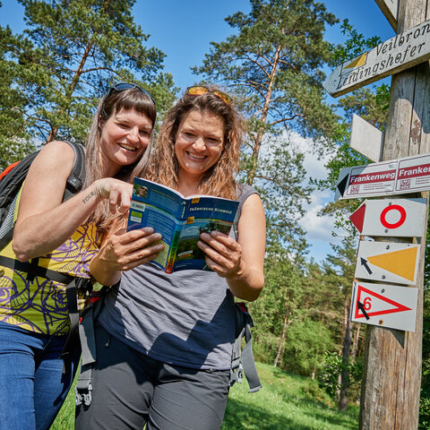 Zwei lachende Frauen stehen an einem Pfosten mit Wanderwegmarkierungszeichen und blättern in einem Reiseführer.