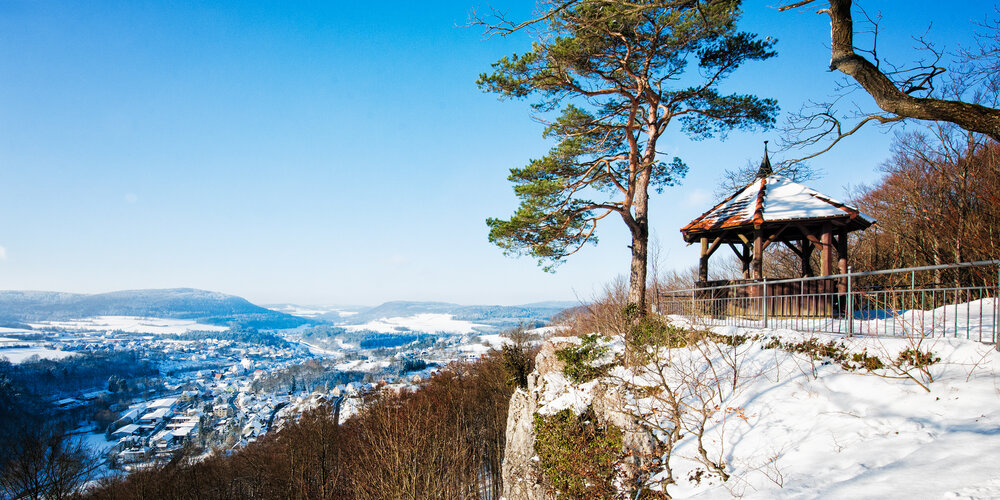 An einer Felskante steht in schneebedeckter Landschaft neben einem Baum ein Holzpavillon. Darunter liegend der Ort.