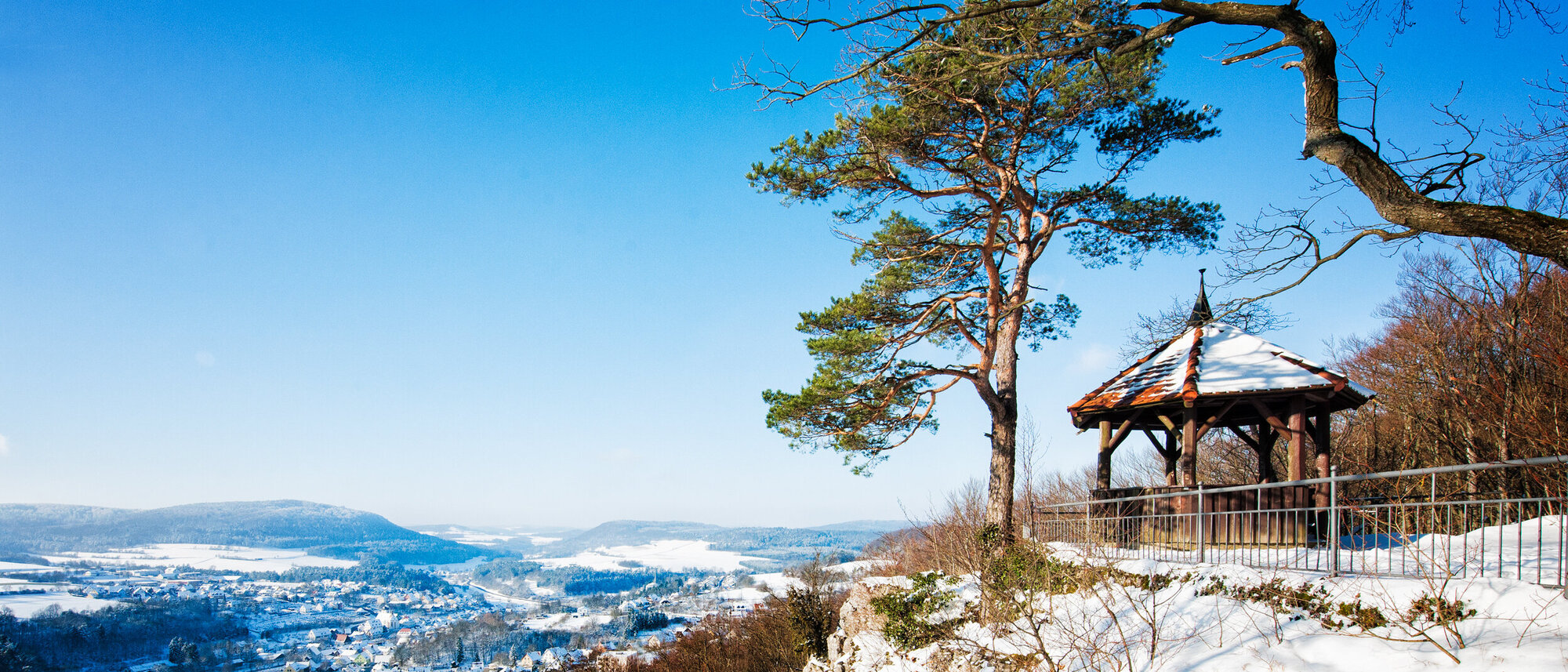 An einer Felskante steht in schneebedeckter Landschaft neben einem Baum ein Holzpavillon. Darunter liegend der Ort.