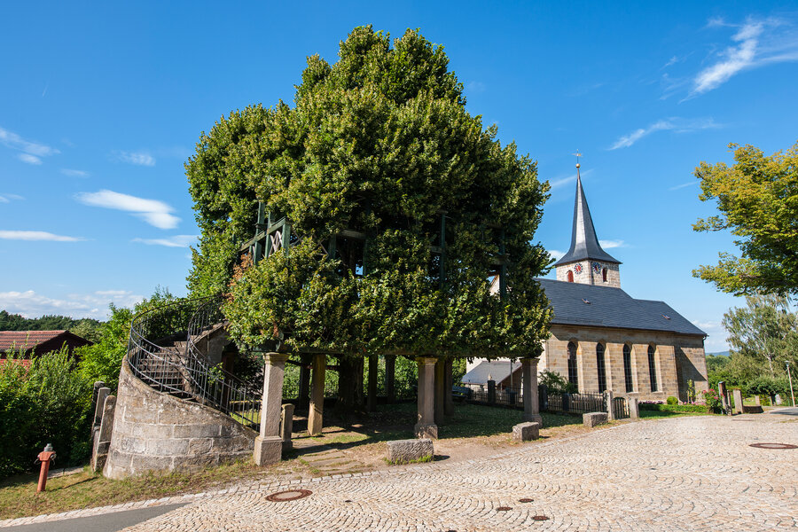 An einem gepflasterten Platz steht eine große Linde, an der eine steinerne Treppe in den Tanzboden führt. Rechts neben der Linde steht eine alte Kirche.