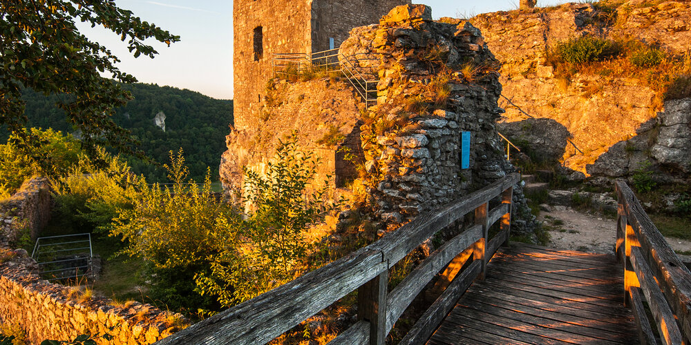 Eine alte Holzbrücke mit Geländer führt hinüber zum Turm der Ruine, die von der Abendsonne angestrahlt wird.