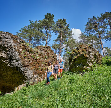 Ein Mann und zwei Frauen wandern lachend über eine Wiese zwischen zwei großen Felsbrocken bergab.
