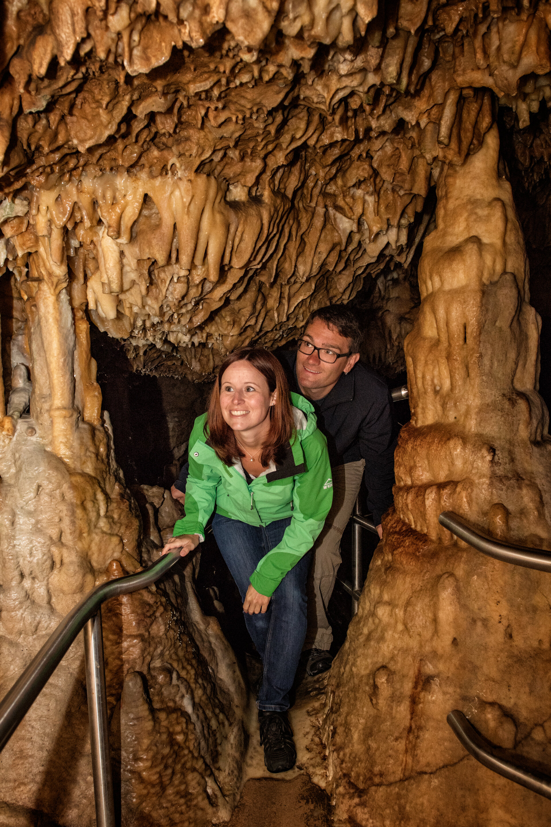 Ein lächelndes Paar läuft geduckt durch eine Höhle. Oben, links und rechts stehen Tropfsteine.