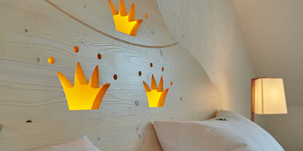 Ein Bett in dessen Kopfteil aus Holz drei Kronen ausgeschnitten sind, die gelb hinterleuchtet werden.