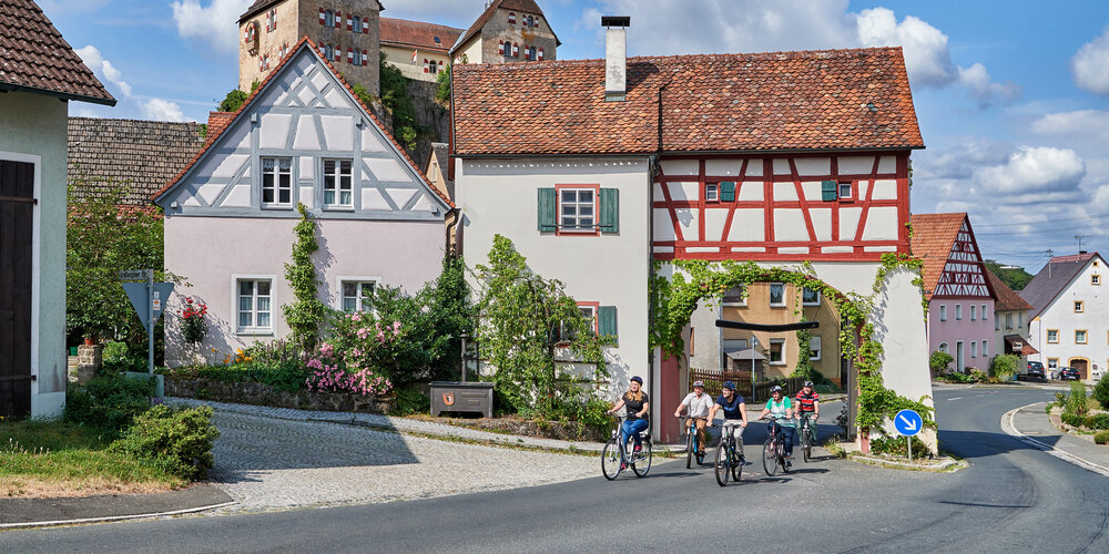Zwei Frauen und drei Männer fahren auf einer Straße durch ein Stadttor mit fränkischem Fachwerk. Im Hintergrund thront eine Burg.