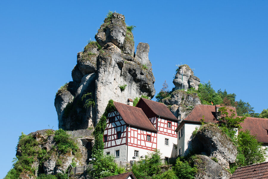 Zwei hochaufragende Felsnadeln, von der eine einen Aussichtspunkt beherbergt. Am Fuße der Felsen klebt ein schönes Fachwerkhaus mit rotem Ziegeldach.