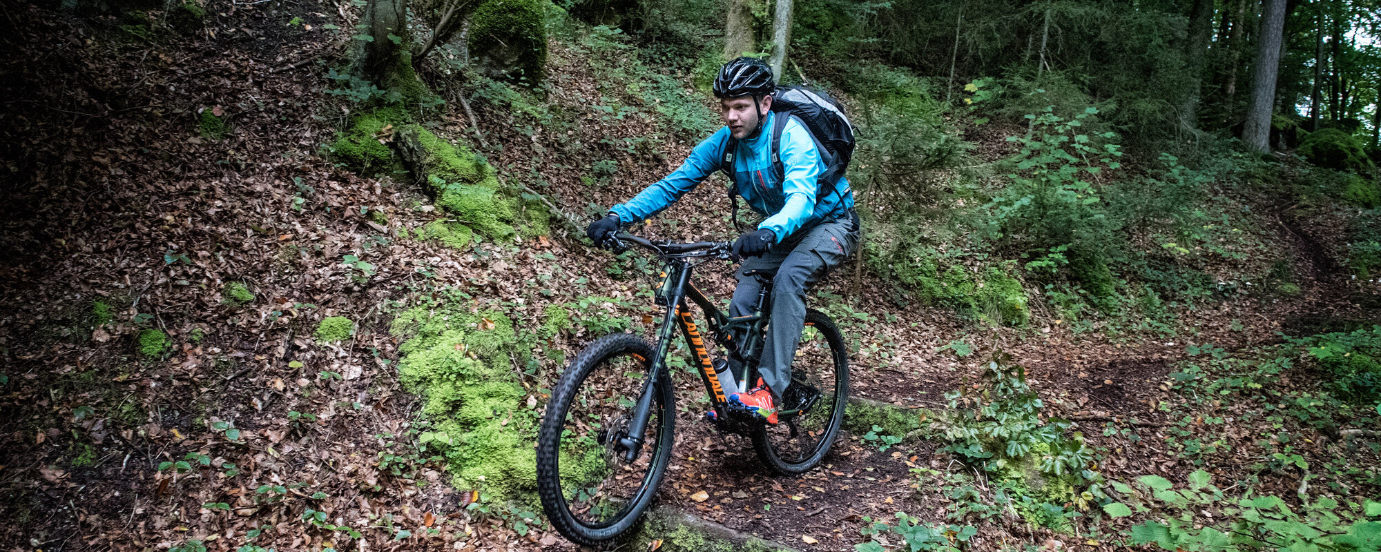 Ein Mann in blauer Jacke sitzt konzentriert auf seinem Fahrrad und fährt auf einem schmalen Pfad im Wald bergab.