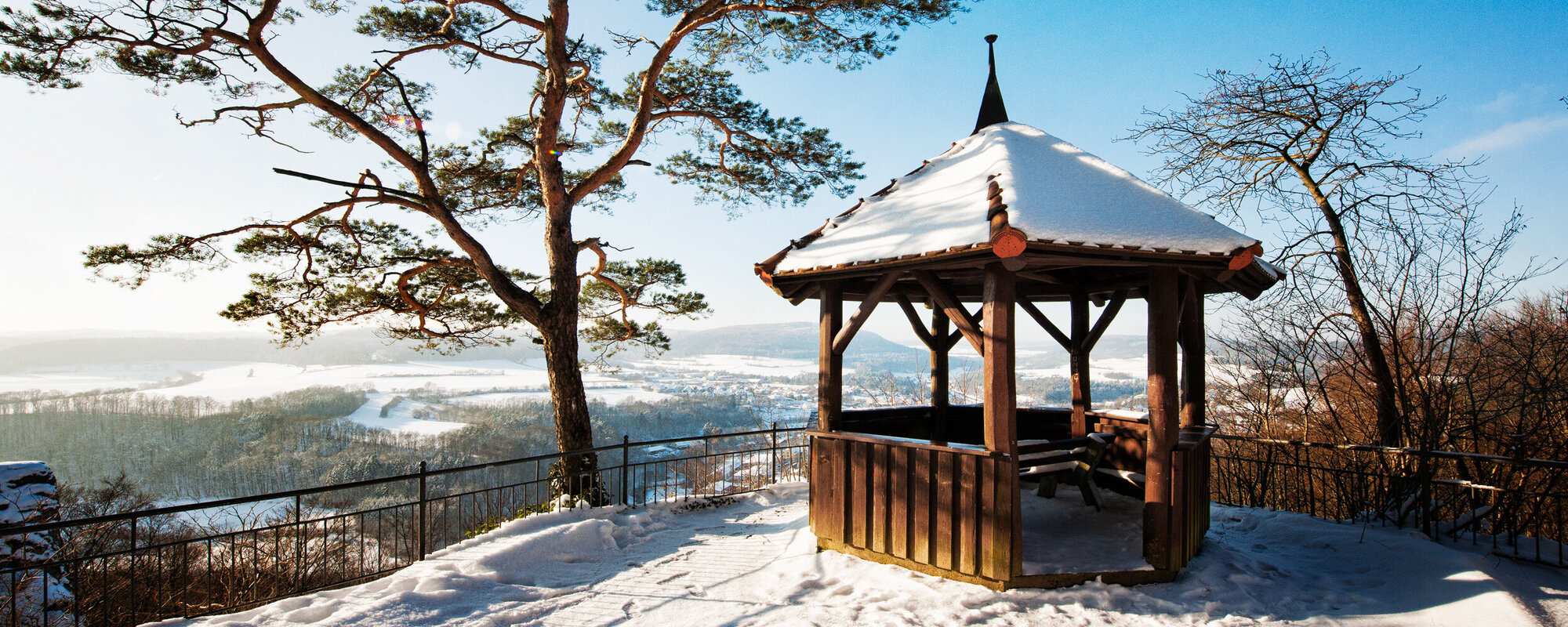 Auf einer Aussichtsplattform steht ein schneebedeckter Holzpavillon neben einem Nadelbaum. Die Plattform ist von einem Metallgeländer eingefasst.