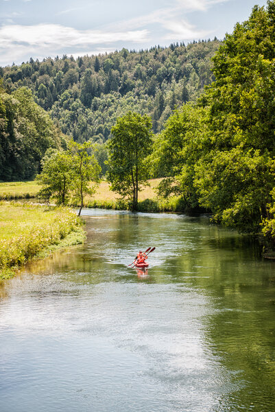 Auf dem Fluss, der von Wiesen und hügeligen Waldern umgeben ist, fahren zwei Personen mit einem roten Kajak.