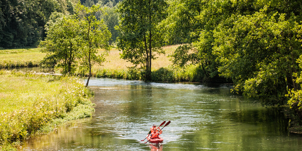 Auf dem Fluss, der von Wiesen und hügeligen Waldern umgeben ist, fahren zwei Personen mit einem roten Kajak.