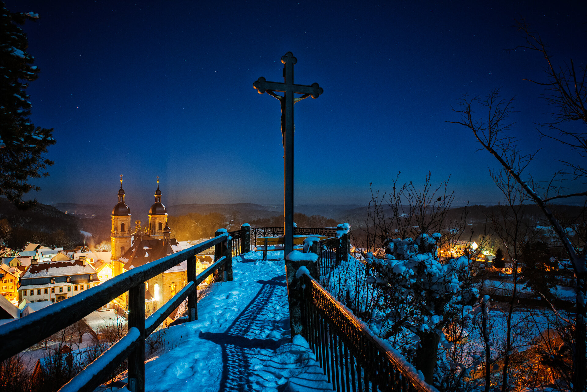 Ein schmaler, verschneiter Weg, der von Geländern eingefasst ist führt zu einem Aussichtspunkt, auf dem ein Keuz steht. Unten, im Hintergrund, steht eine Kirche. Der dunkelblaue Himmel ist sternenklar.