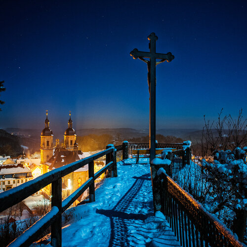 Ein schmaler, verschneiter Weg, der von Geländern eingefasst ist führt zu einem Aussichtspunkt, auf dem ein Keuz steht. Unten, im Hintergrund, steht eine Kirche. Der dunkelblaue Himmel ist sternenklar.
