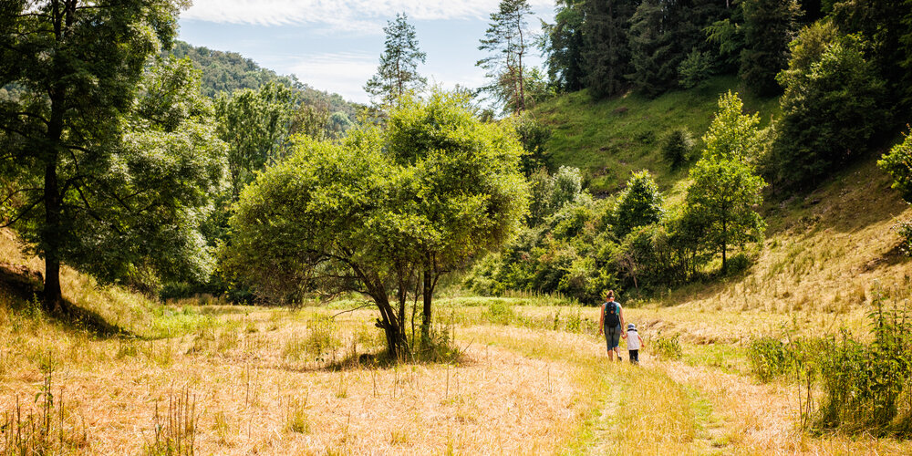 Eine Frau wandert mit ihrem Kind durch ein Trockental, das links und rechts mit Bäumen gesäumt ist.