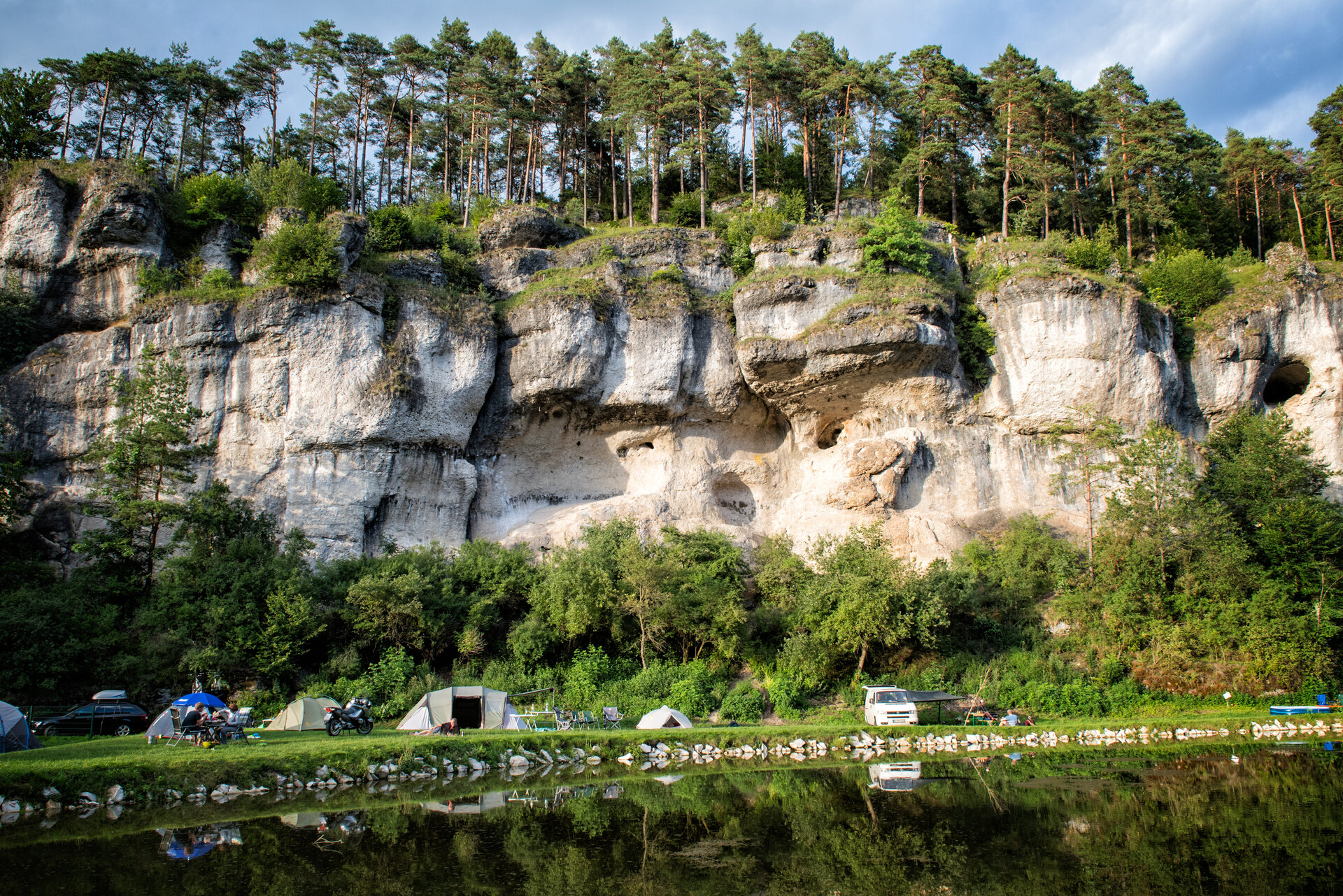 Eine Breite Felswand und der darunter liegende Campingplatz spiegeln sich in einem Weiher.