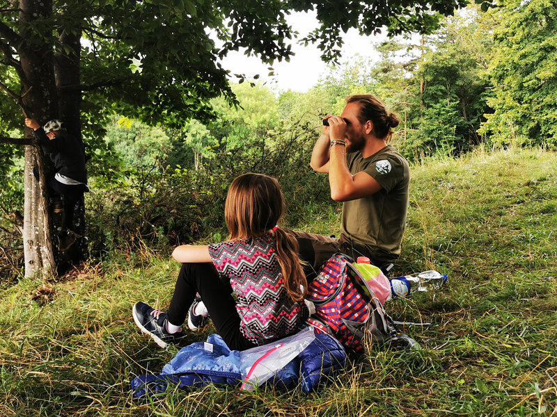 Ein Naturparkranger, der durch ein Fernglas blickt, sitzt mit einem Mädchen auf einer Wiese.