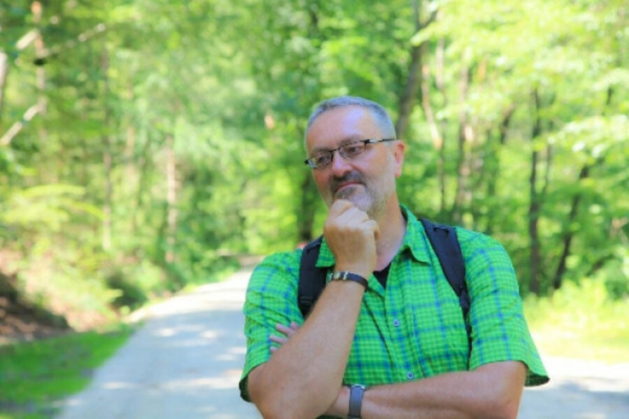 Ein Mann im grünen Hemd steht lächelnd auf einem Feldweg vor einem Wald.