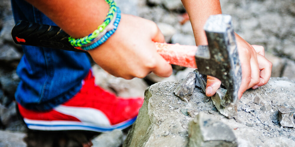 Zwei Kinderhände mit einem Hammer in der Hand, mit dem auf einen Stein geklopft wird.