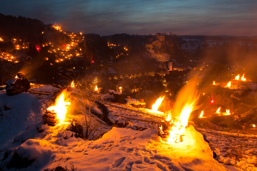 Im Vordergrund stehen mehrere brennende Holzstapel bei Einbruch der Nacht oberhalb der Stadt. Im Hintergrund sind weitere Feuerstellen und eine Burg zu sehen.