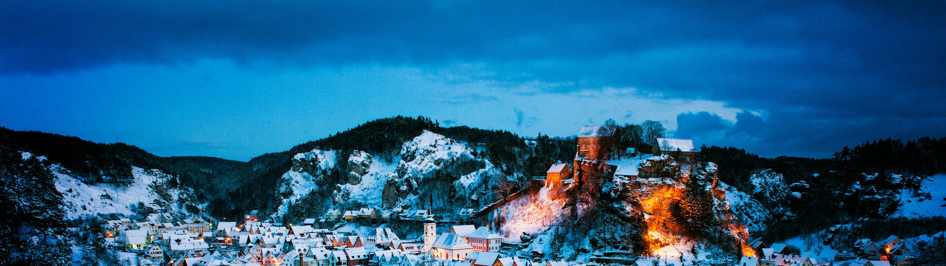 Blick von oben auf die verschneiten Dächer und beleuchteten Straßen. Auf einem Felsen erhebt sich die Burg.