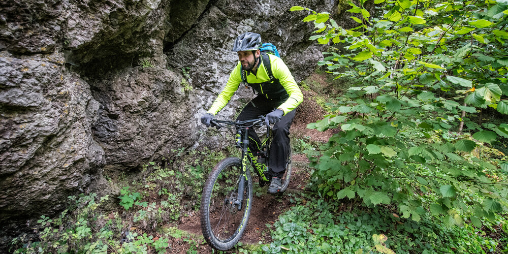 Ein konzentrierter Radfahrer fährt auf einem schmalen Waldpfad direkt an einer grauen Felswand entlang.