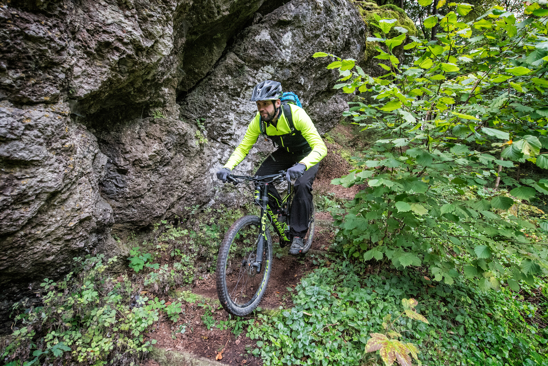 Ein konzentrierter Radfahrer fährt auf einem schmalen Waldpfad direkt an einer grauen Felswand entlang.