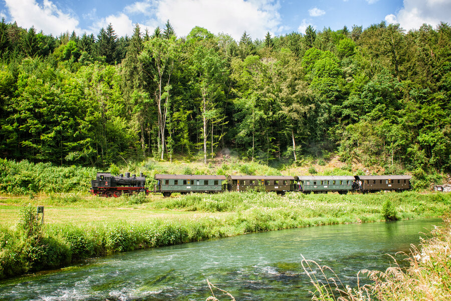 Eine alte Eisenbahn mit vier Waggons fährt durch ein Tal. Im Vordergrund ist ein Fluss.
