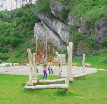 Vor einer grauen Felswand ist ein großes rotes Netz für Kinder zum Klettern und ein Parcours aus Holzstämmen zum Balancieren.