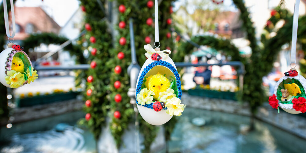 In einem aufgeschnittenen Ei sitzt ein kleines Kunststoffküken. Das Ei hängt an einer Schnur am Osterbrunnen, der im Hintergrund zu erahnen ist.