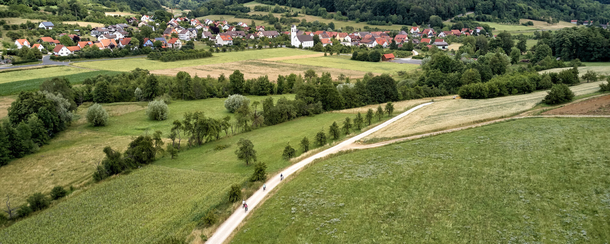 Im Vordergrund fahren vier Radfahrer auf einem Feldweg. Im Hintergrund ist ein Ort in Tallage vor bewaldeten Hügeln zu sehen.