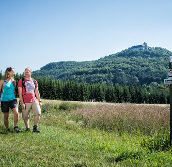 Ein paar wandert lächelnd auf einem Weg über eine Wiese. Im Hintergrund thront das Schloss auf einem bewaldeten Berg.