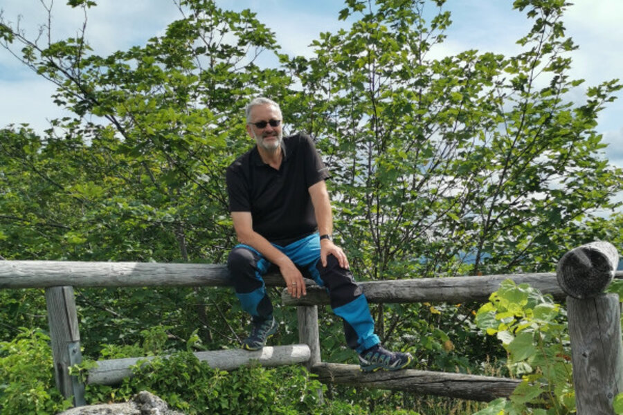 Ein Mann sitzt auf einem Holzgeländer vor einem Busch und trägt eine Sonnenbrille.