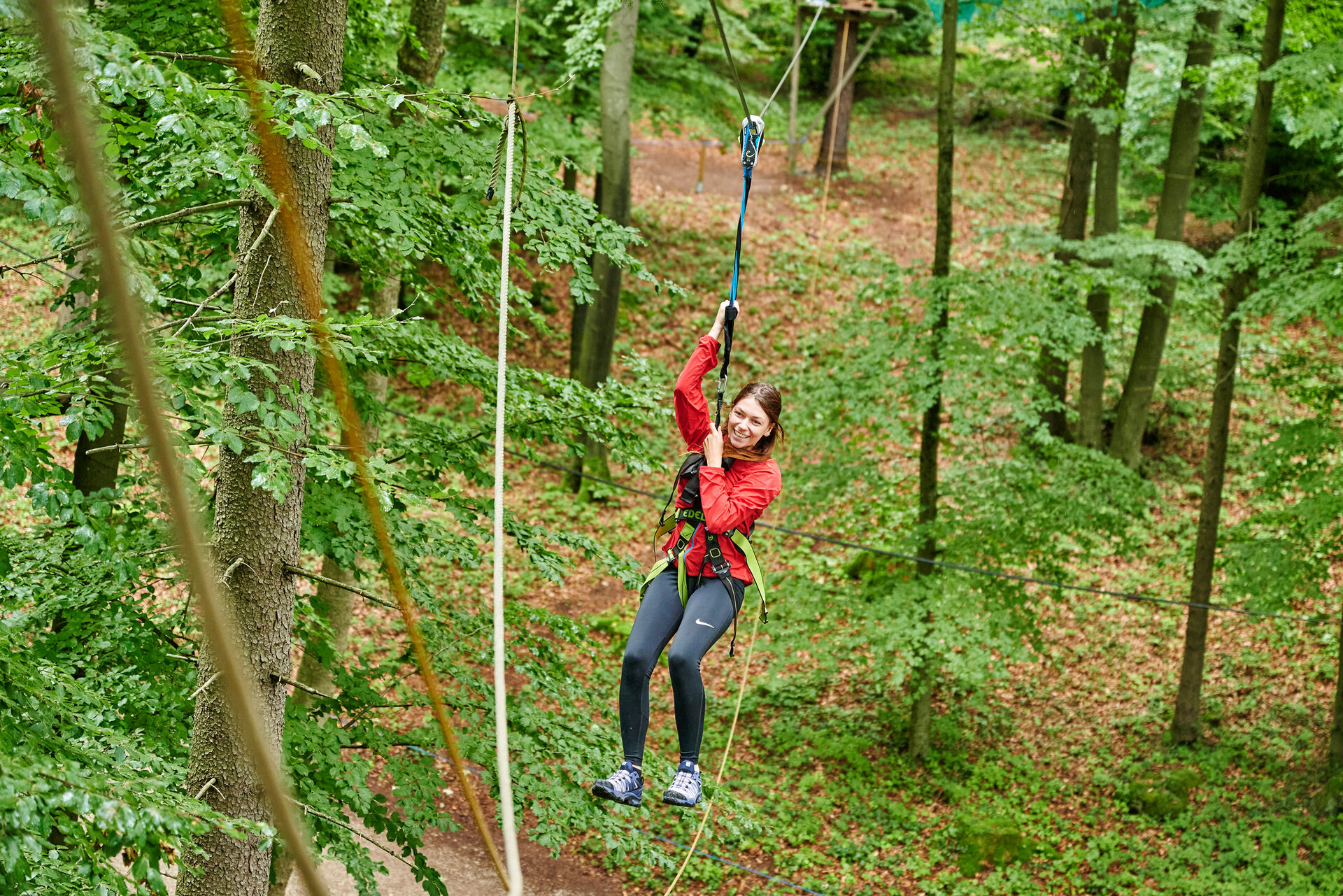 Eine Frau in roter Jacke und schwarzer Hose hängt an einem gesichterten Seil und schwebt von einem Baum zum anderen.