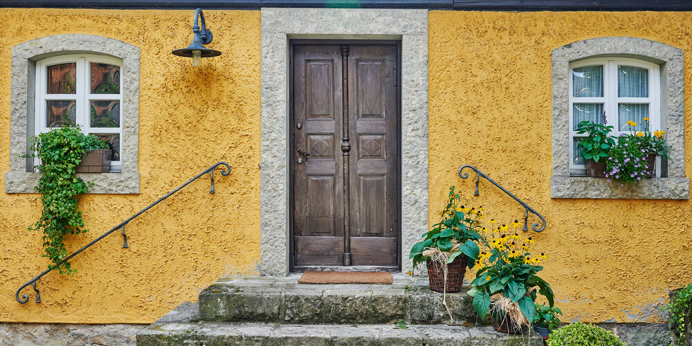 Eine alte hölzerne Eingangstür führt über Steintreppen in ein gelbes Haus.