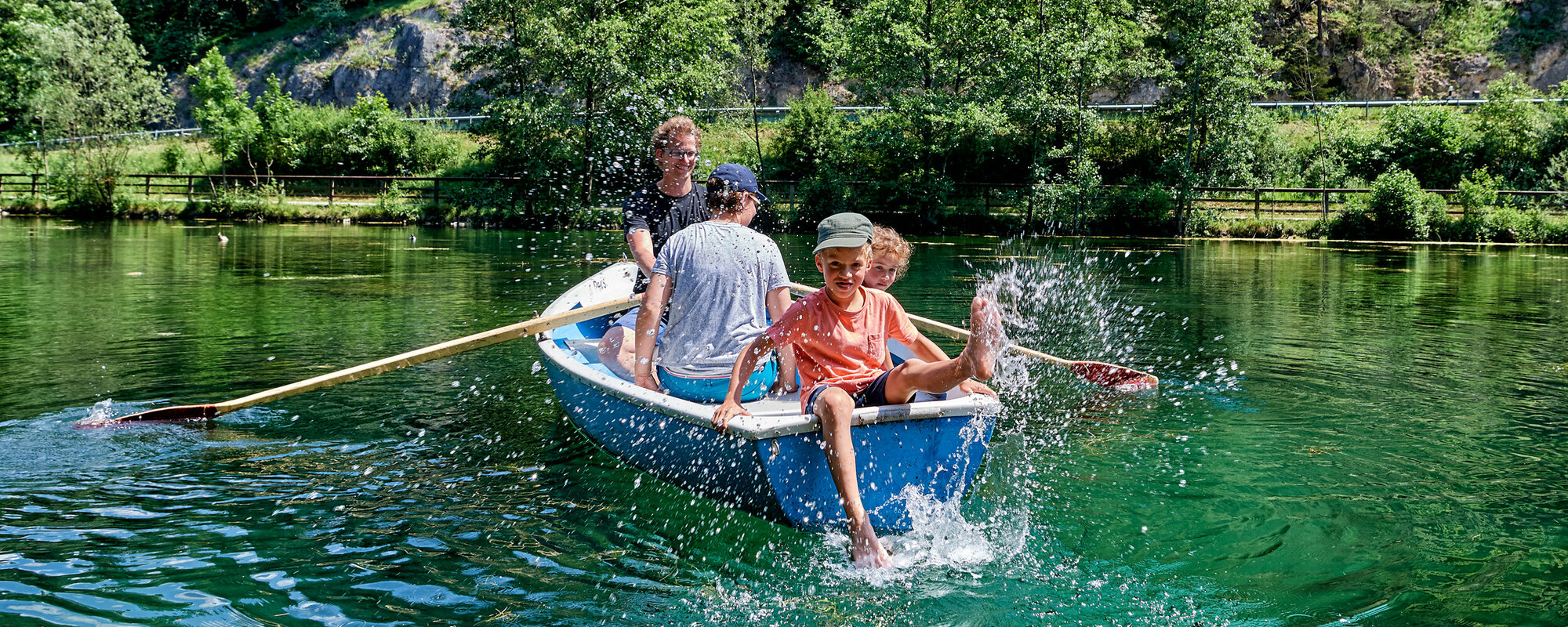 Eine Familie sitzt in einem blauen Boot auf einem See, der von Wald umgeben ist. Der Vater rudert, der Sohn sitzt hinten an der Kante und spritzt mit dem Fuß mit Wasser.