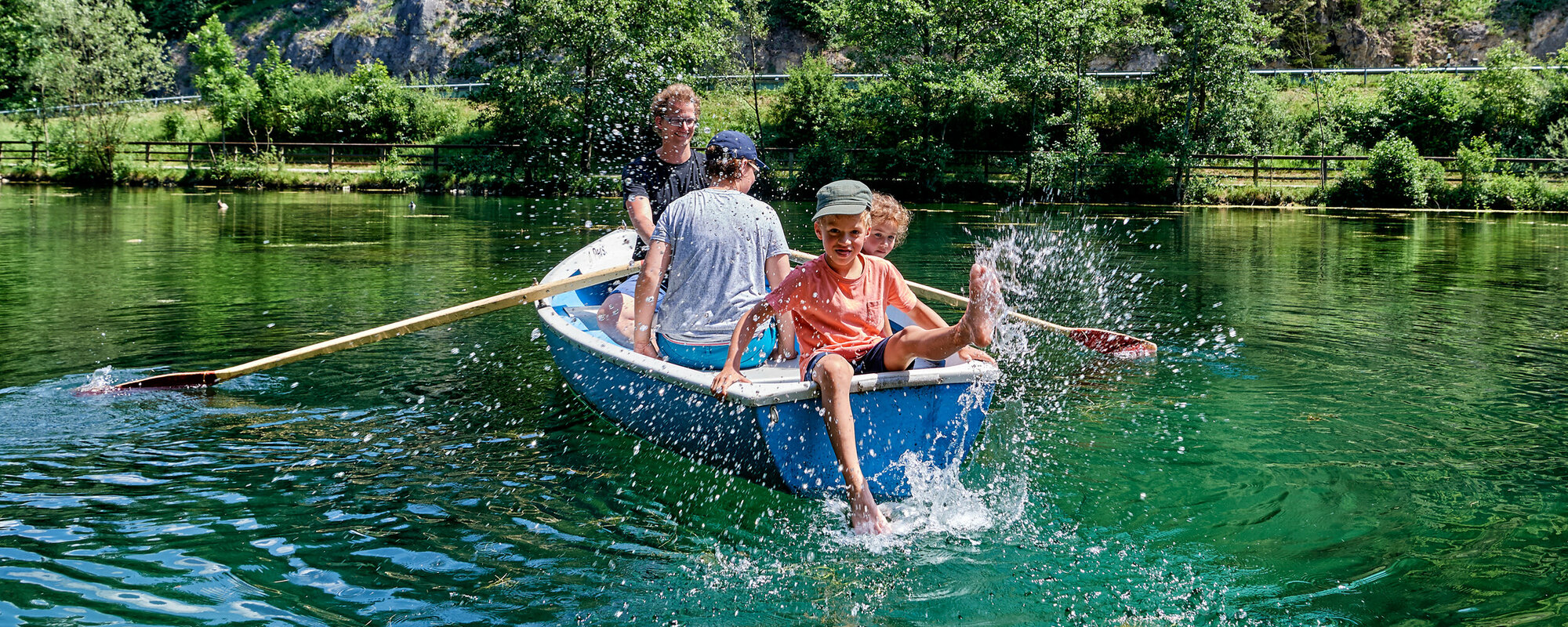 Eine Familie sitzt in einem blauen Boot auf einem See, der von Wald umgeben ist. Der Vater rudert, der Sohn sitzt hinten an der Kante und spritzt mit dem Fuß mit Wasser.