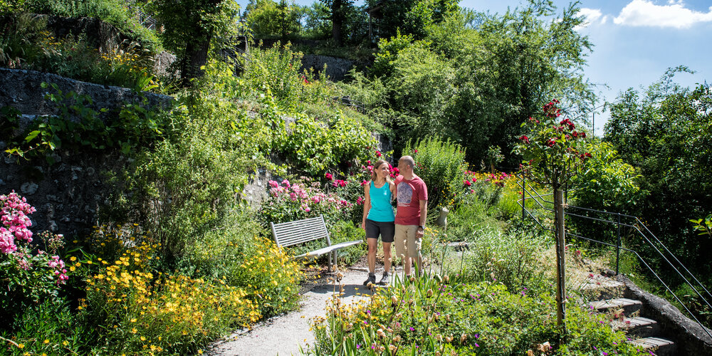 Ein Mann und eine Frau laufen lächelnd durch eine Gartenanlage mit vielen bunten Blumen.